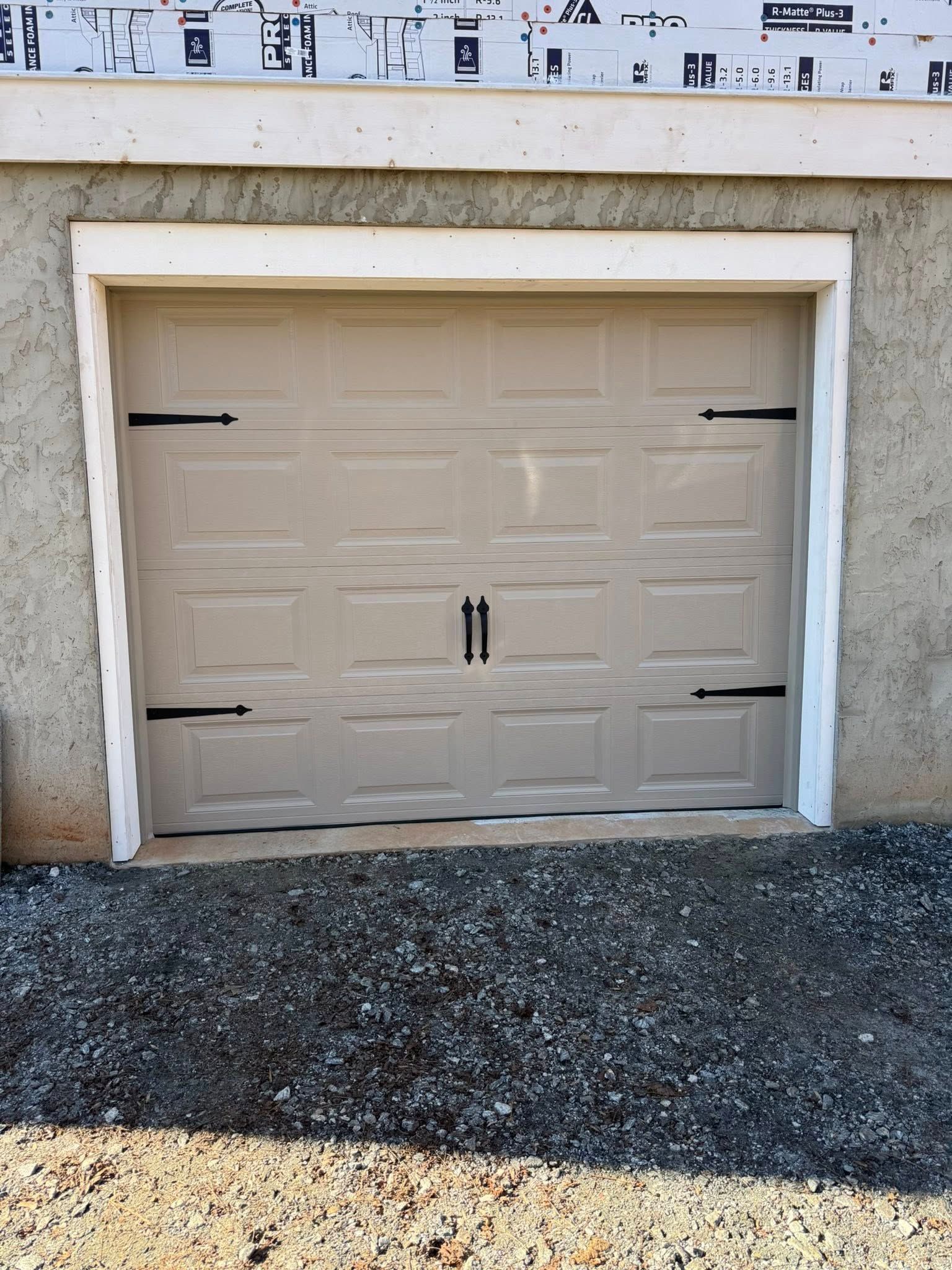 Beige garage door with black handles and tape marks, set in a stucco wall beside gravel.