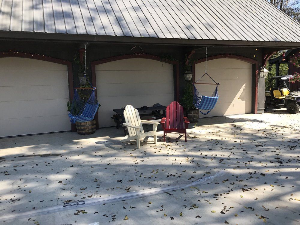 Two chairs on a sunny driveway in front of garage doors, with hanging baskets nearby