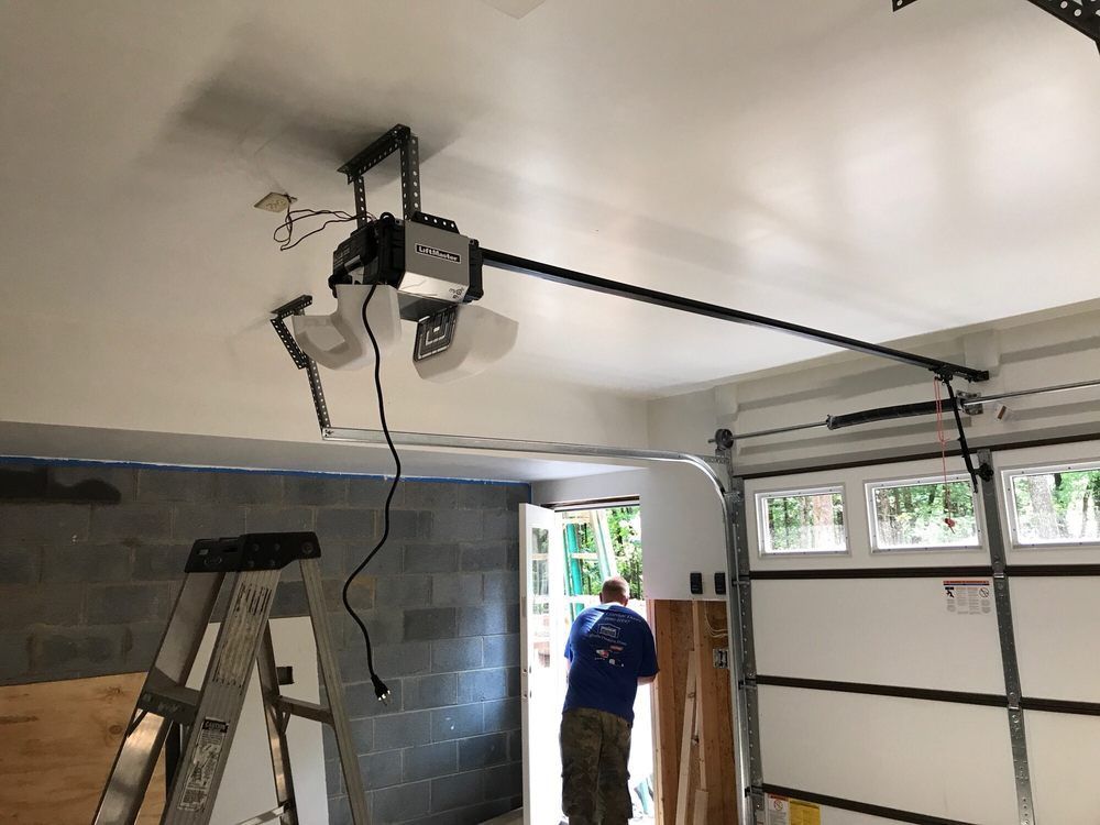Person in a garage standing near an open door, with a ladder and ceiling-mounted garage door opener visible