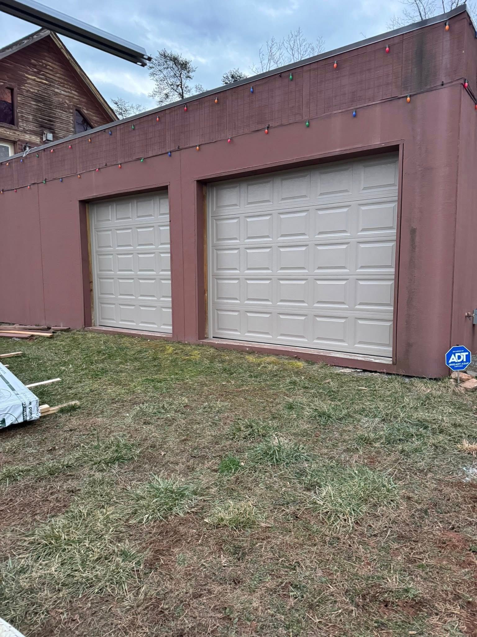 Two beige garage doors on a maroon outbuilding, with a grassy yard in front.