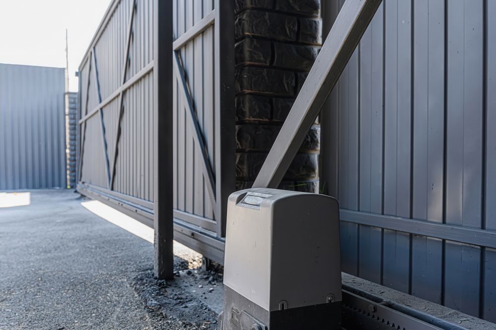 Sliding gray metal gate beside a blue fence, with a driveway opener box at the base