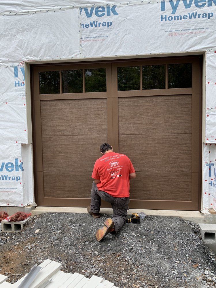 Worker kneeling at a garage entrance, installing brown double garage doors under house wrap.