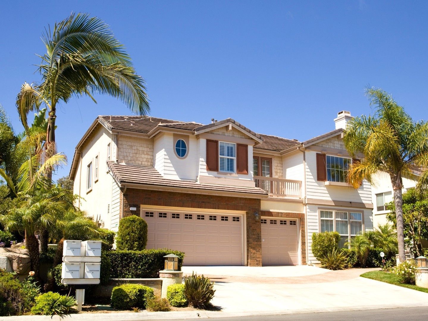 Suburban two-story beige house with palm trees, a garage, and a clear blue sky