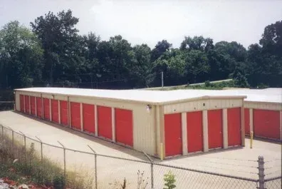 Row of red storage unit doors in a beige building beside a chain-link fence and trees