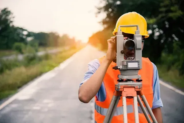 un homme en gilet de sécurité orange et casque jaune regarde à travers un télescope sur une route .