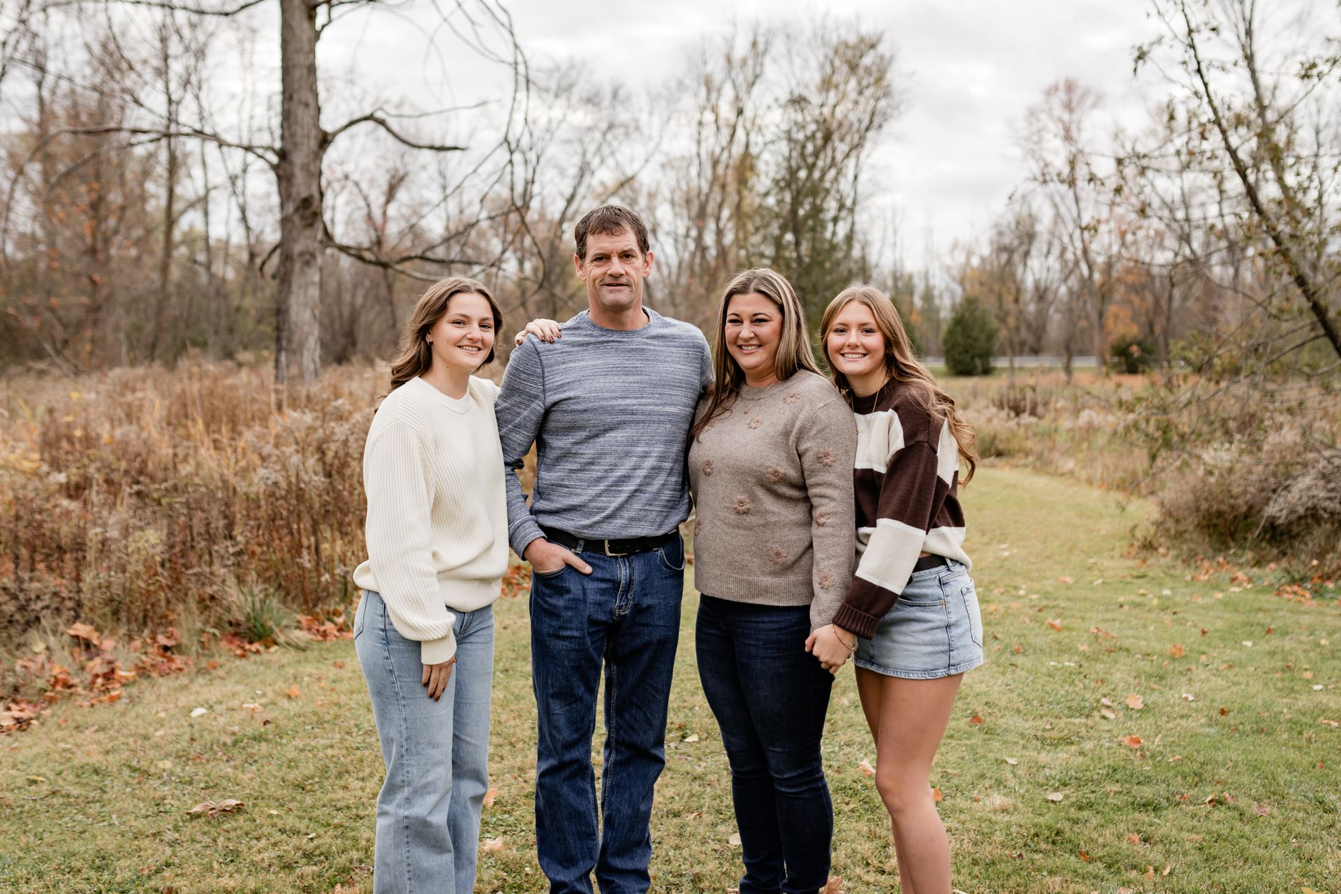 Four people pose standing side-by-side in a grassy field with trees in the background on an autumn day.