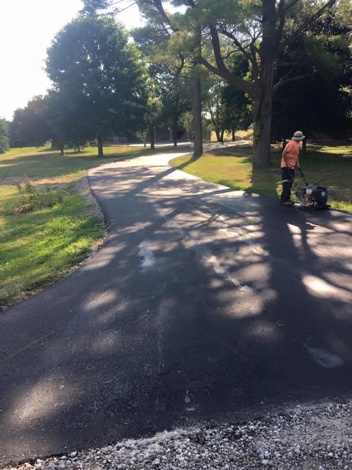 A construction worker operates a plate compactor on a newly paved asphalt driveway in a wooded, grassy area.