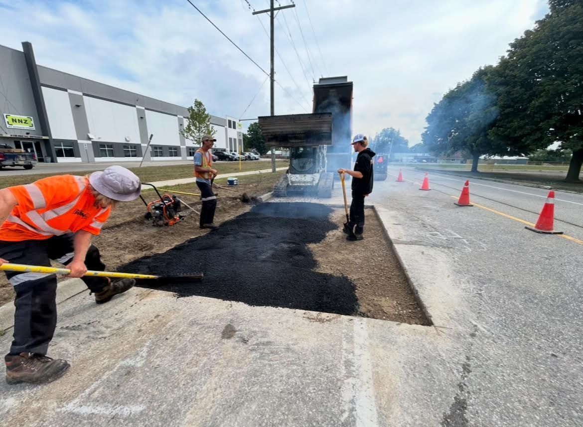 Road crew workers in high-visibility gear paving a street with asphalt near a warehouse.