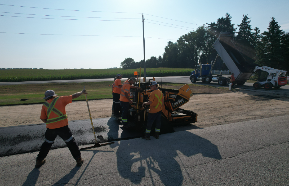 Road crew in safety gear patching asphalt on a road with machinery and a dump truck in the background.