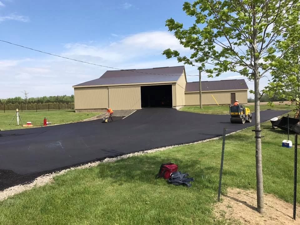 Construction workers use a small steamroller on a newly paved black asphalt driveway in front of a tan pole barn.