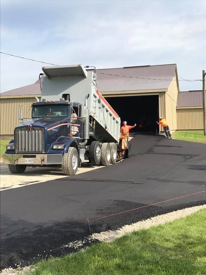 A dump truck pours asphalt to pave a driveway in front of a metal building, with workers guiding the process.