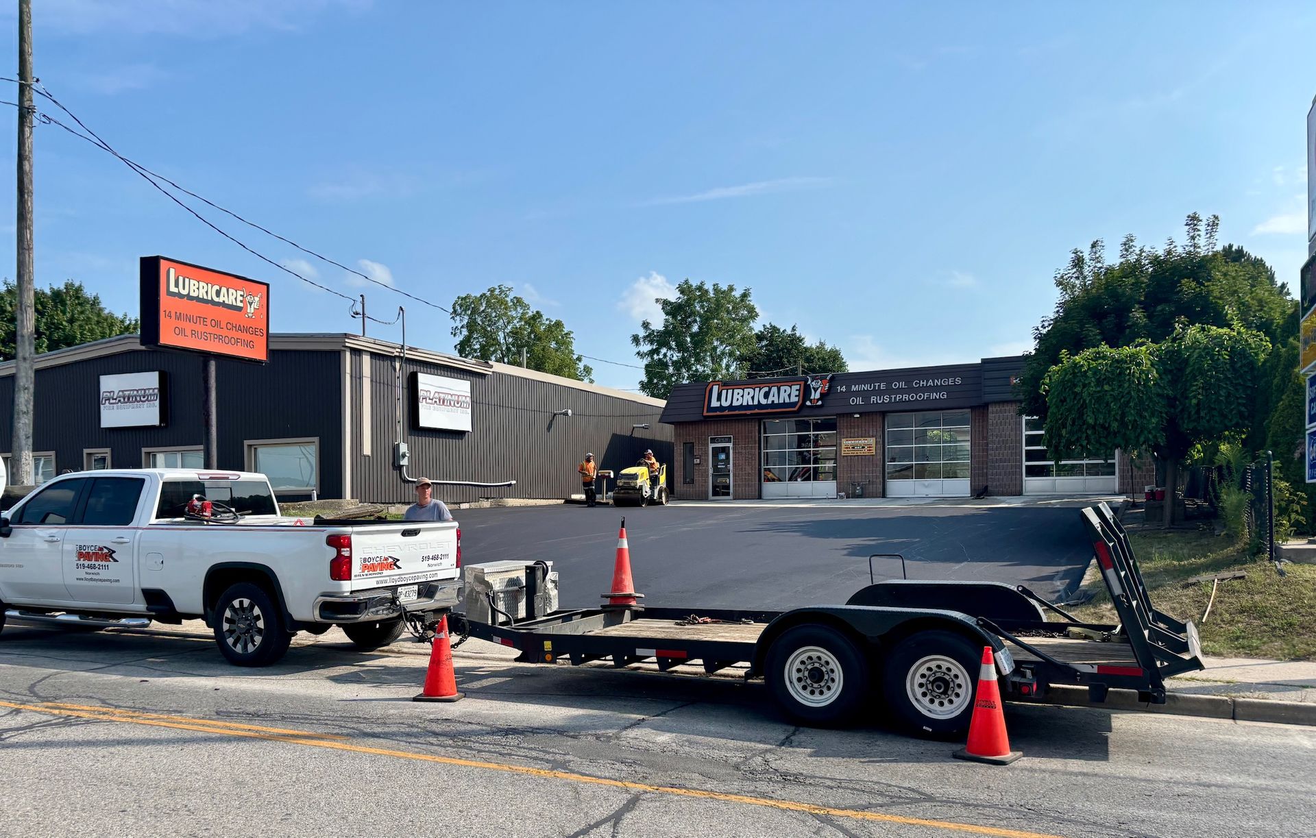 A white pickup truck with an empty flatbed trailer parked in front of a newly paved asphalt parking lot at a business.