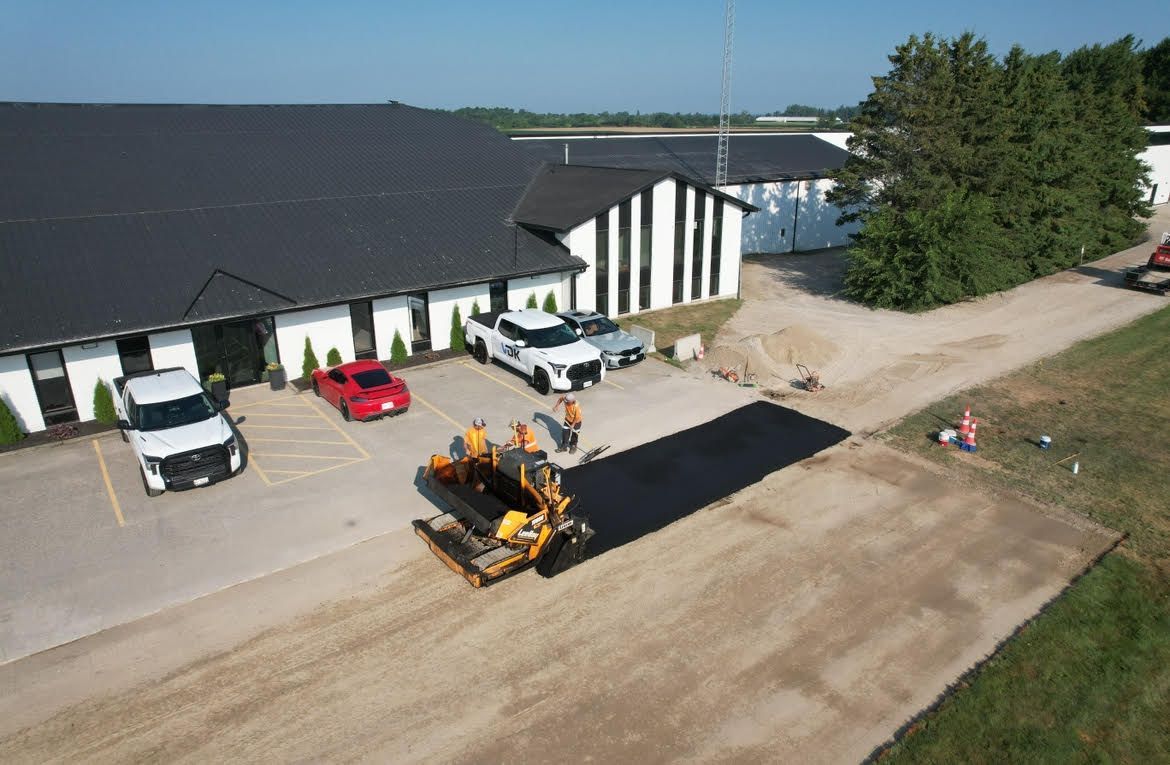 An aerial view shows workers paving a driveway with black asphalt in front of a modern white building with a black roof.