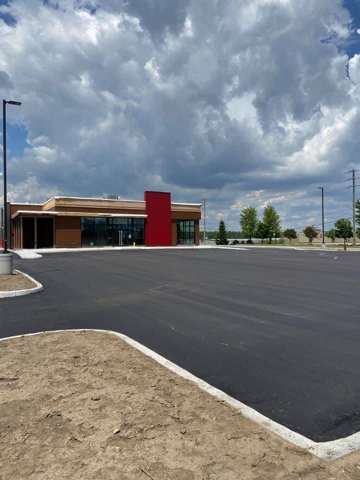 A newly paved parking lot leads to a modern commercial building with tan siding and a prominent bright red entrance pillar.