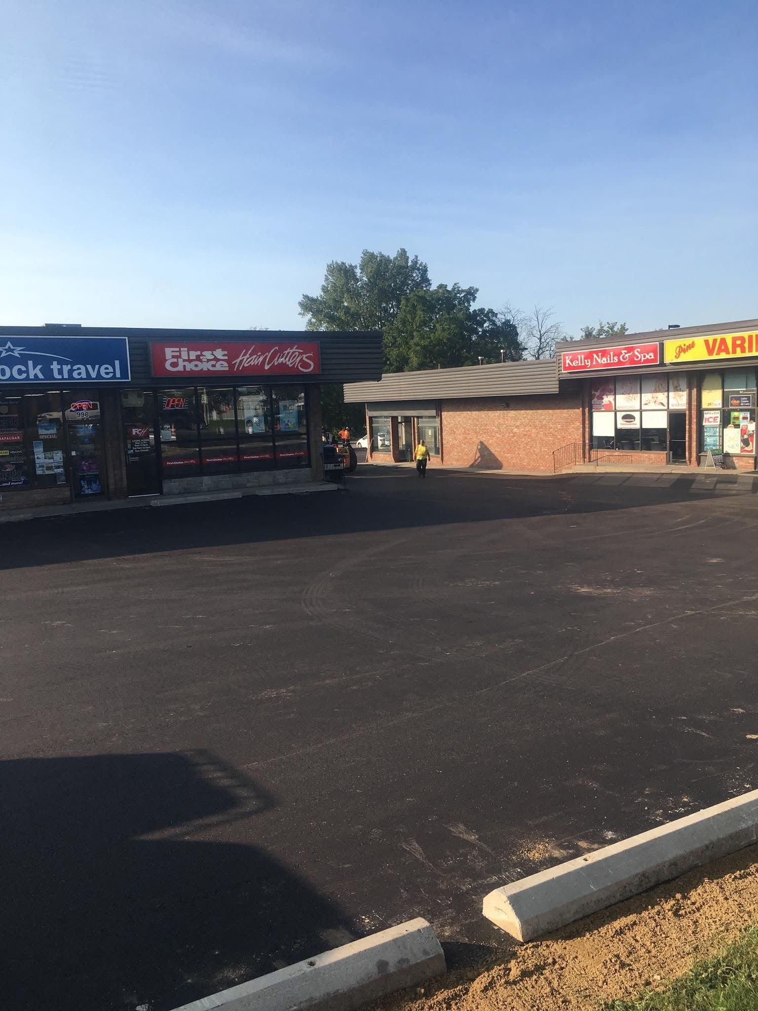 A strip mall exterior with storefronts, including a travel agency and a restaurant, viewed across a large, paved lot.