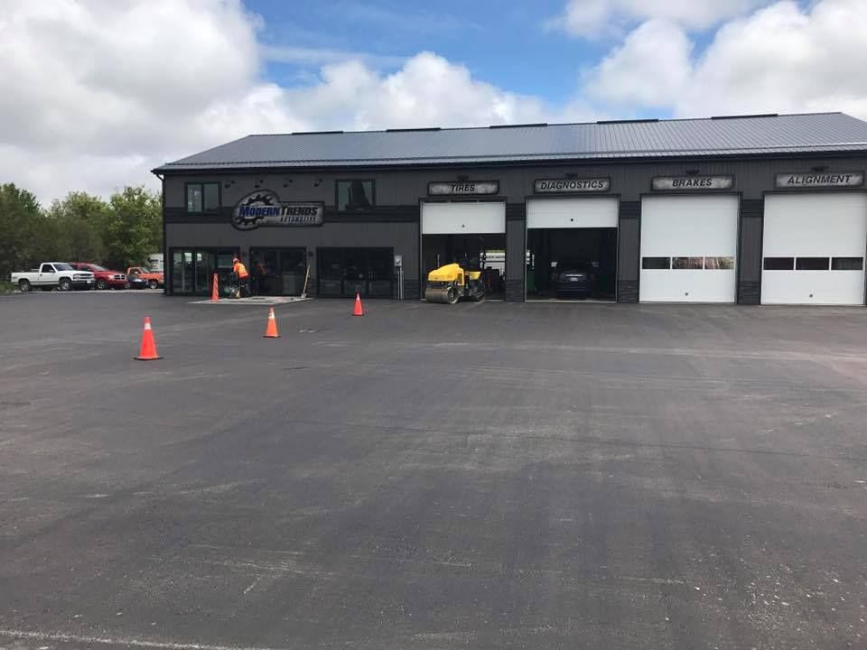 A dark-grey commercial service building with four garage bays, an asphalt parking lot, and traffic cones.