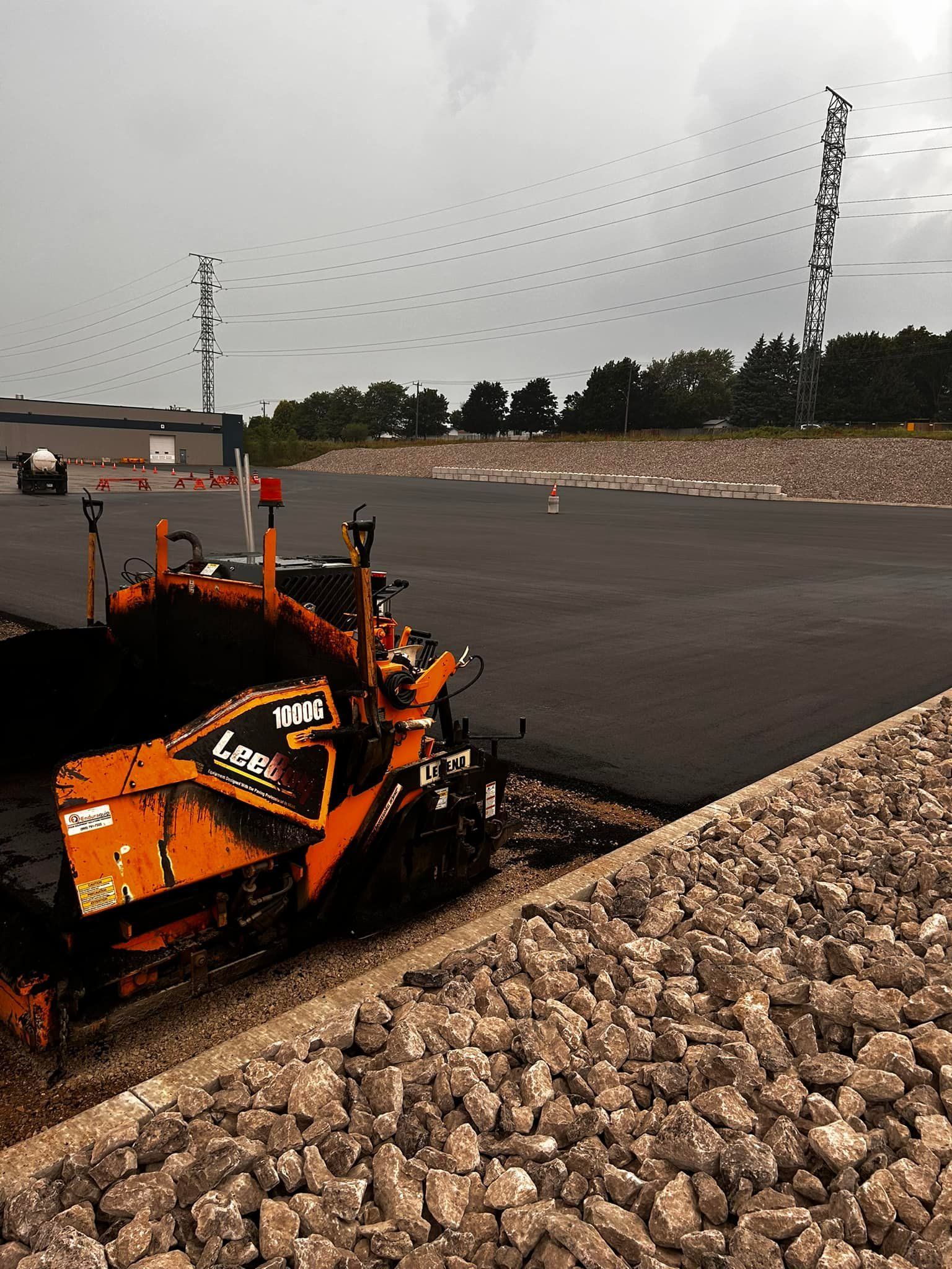 An orange asphalt paver works on a large, freshly paved road surface next to a rock-covered embankment.