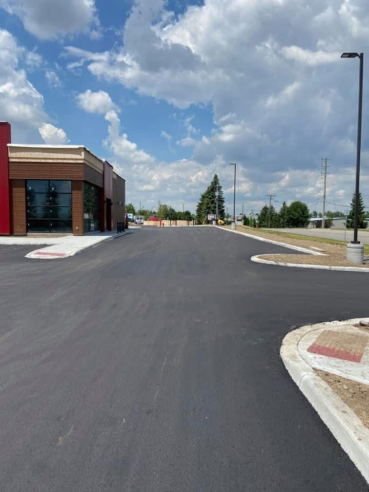 Newly paved asphalt road and parking area next to a brick commercial building under a blue sky with white clouds.