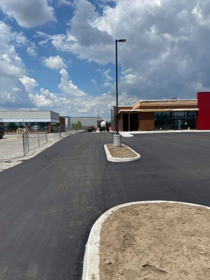 Freshly paved asphalt parking lot with concrete curbs and landscaping under a blue sky with clouds near a commercial building.
