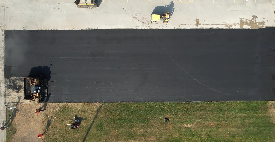An aerial view of a freshly paved black asphalt driveway patch bordered by concrete and green grass.