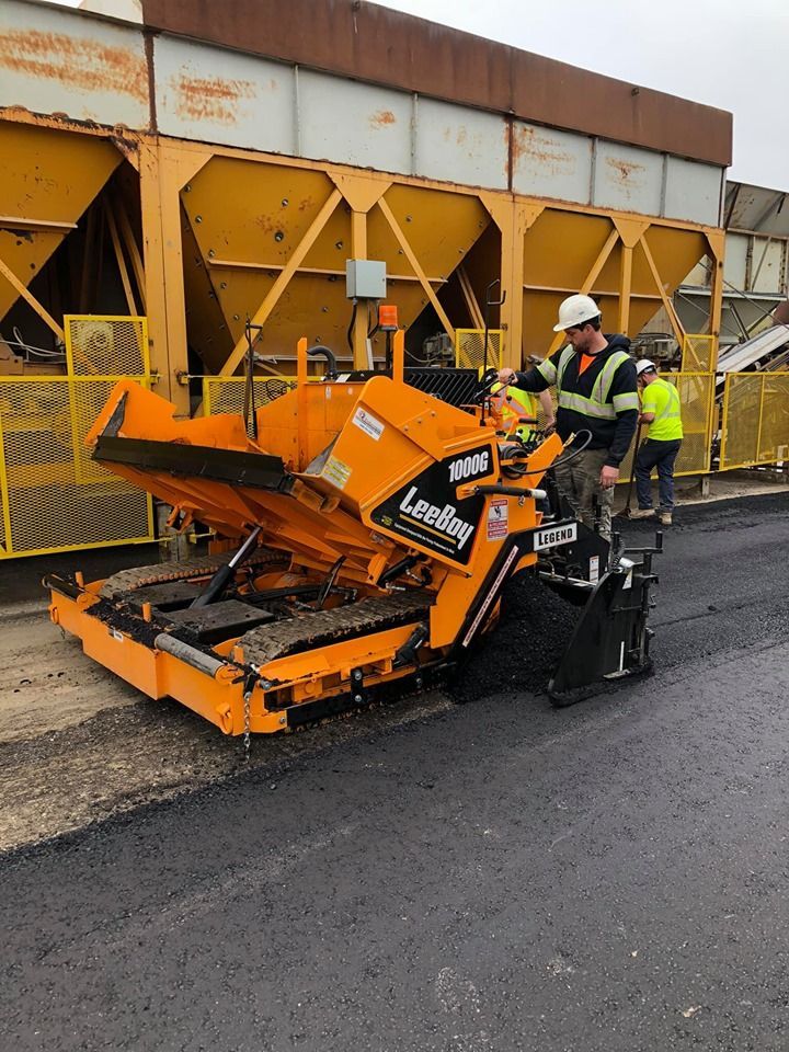 A worker operates an orange LeeBoy asphalt paver in front of large industrial storage bins.