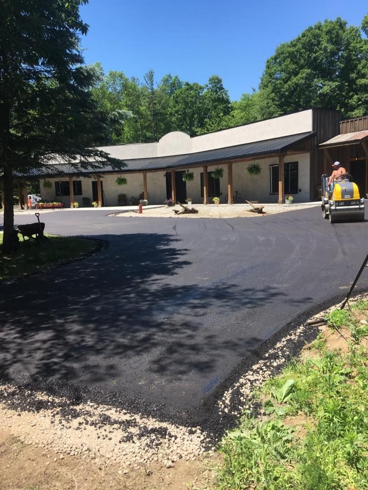 A construction worker on a yellow steamroller paves a dark asphalt lot in front of a modern building with a porch.
