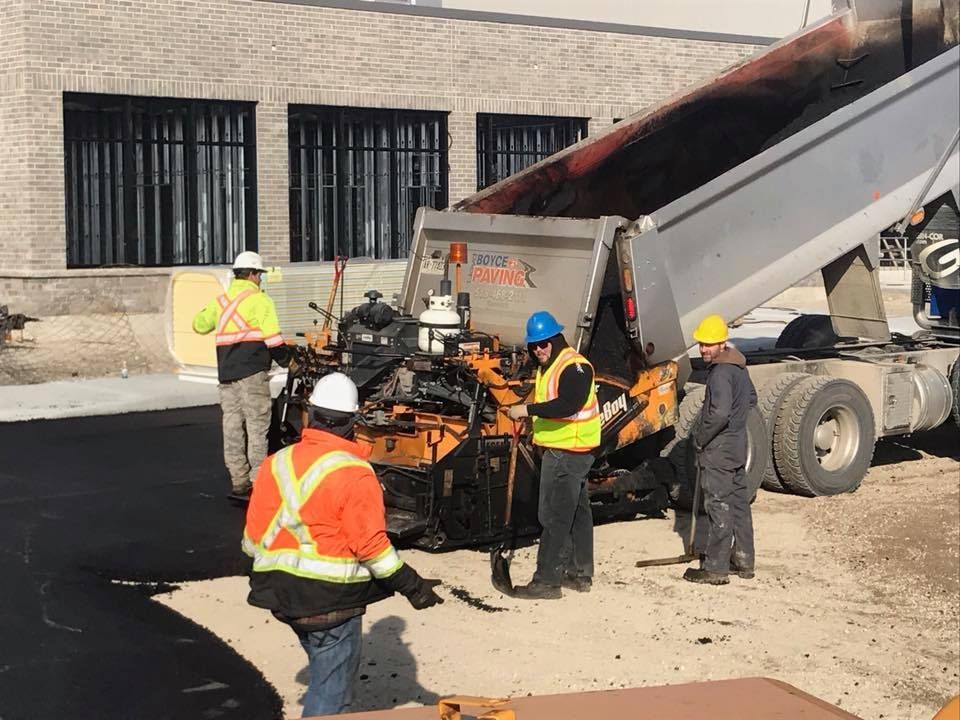 Workers in high-visibility vests operate a paving machine alongside a dump truck at a construction site.