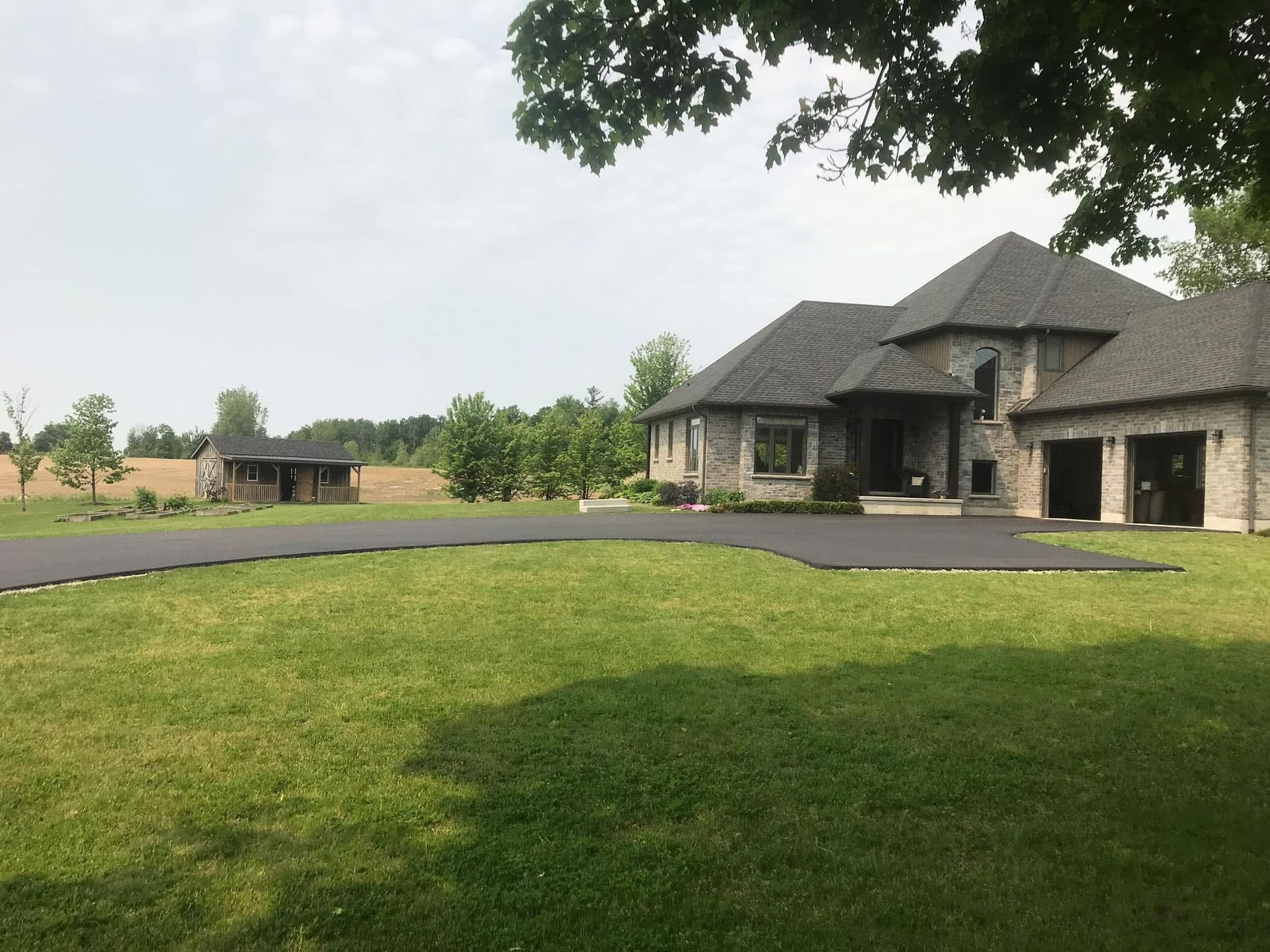 A large stone house with a multi-car garage, a wide paved driveway, and a small outbuilding in a grassy, rural setting.