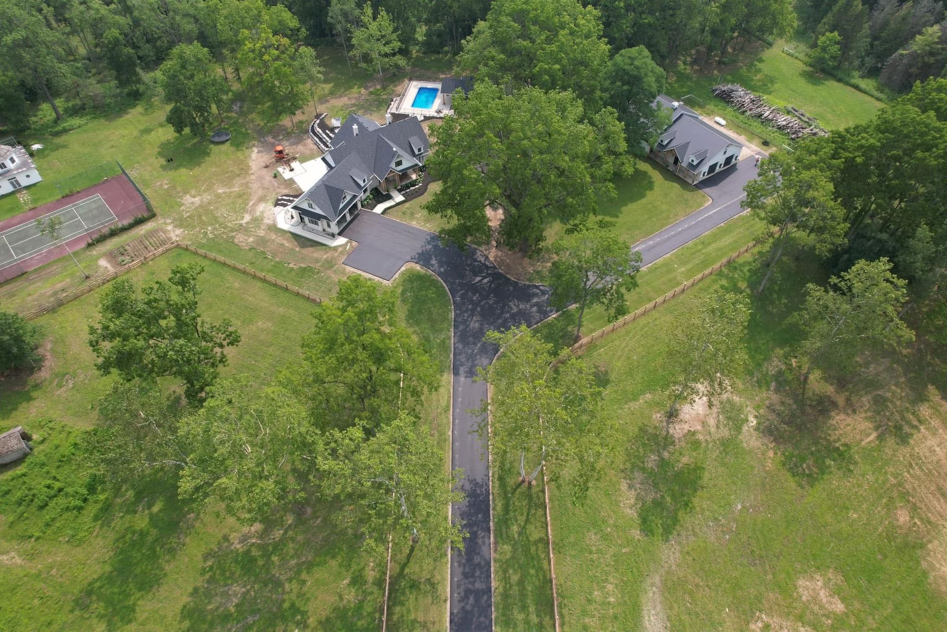 Aerial view of two dark-roofed houses, a pool, and a tennis court situated on a large, tree-filled grassy property.
