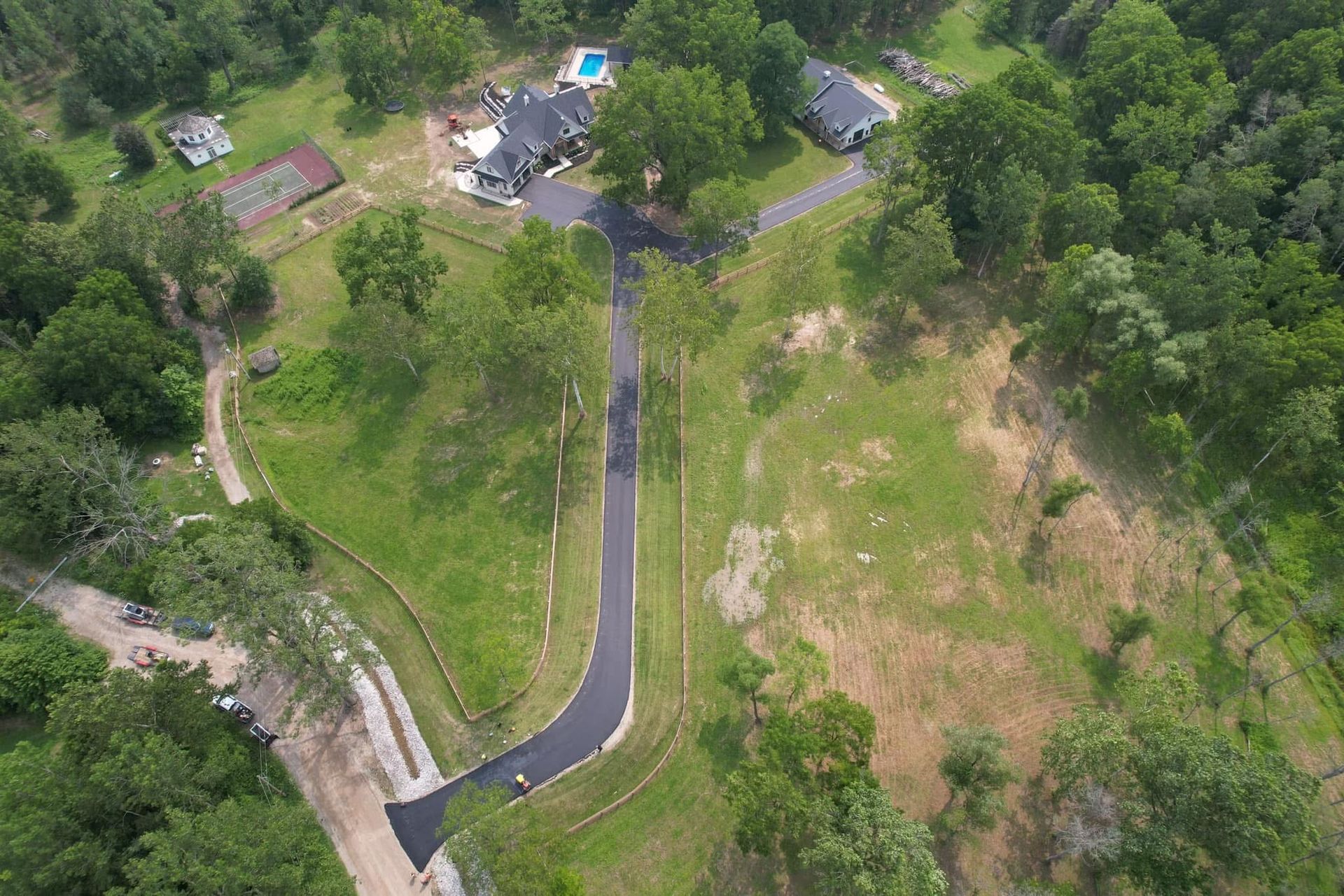 Aerial view of a paved driveway leading to a house with a pool, surrounded by green lawns and dense trees.