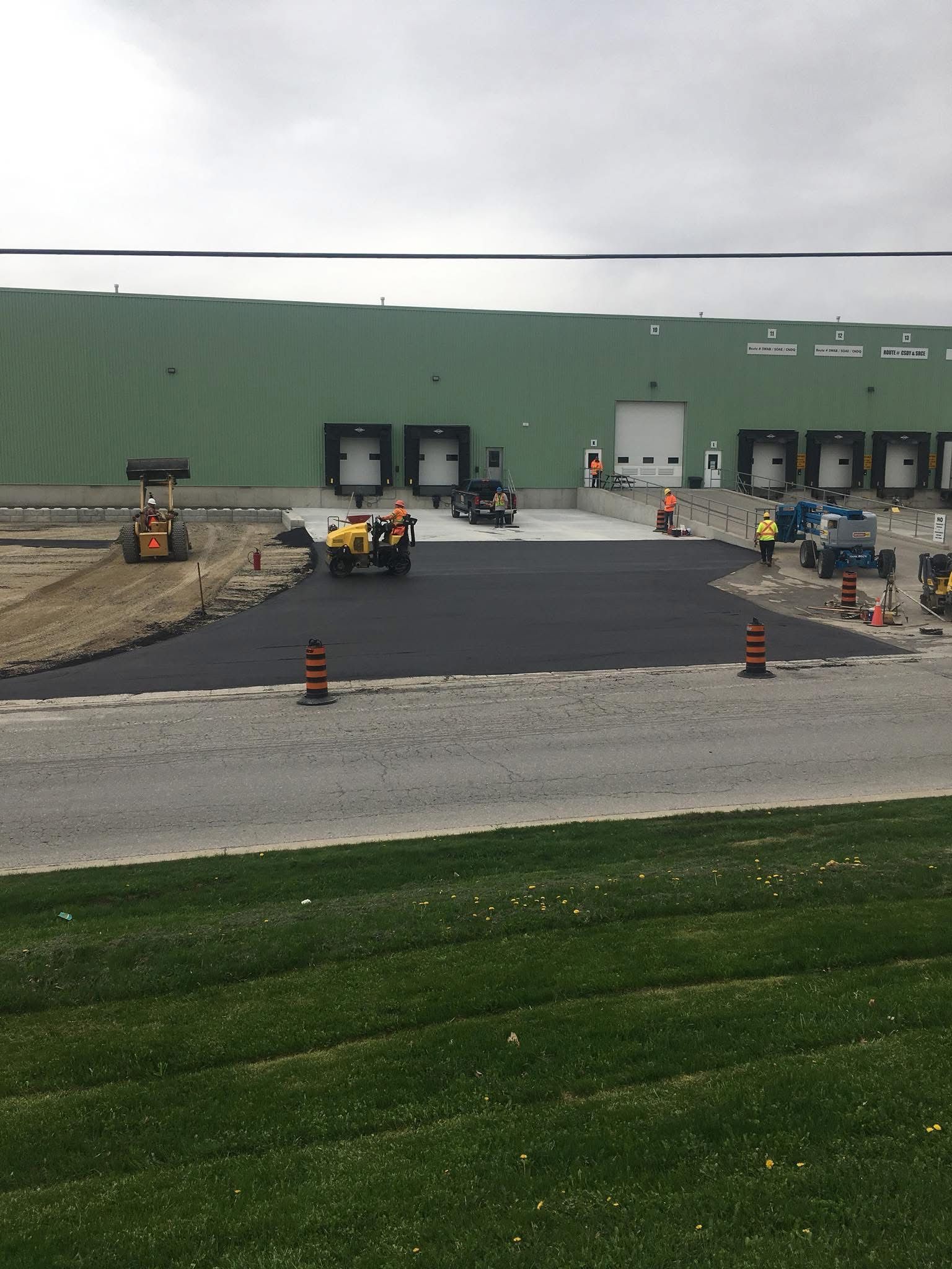 Construction workers paving a driveway at an industrial warehouse with loading docks under an overcast sky.