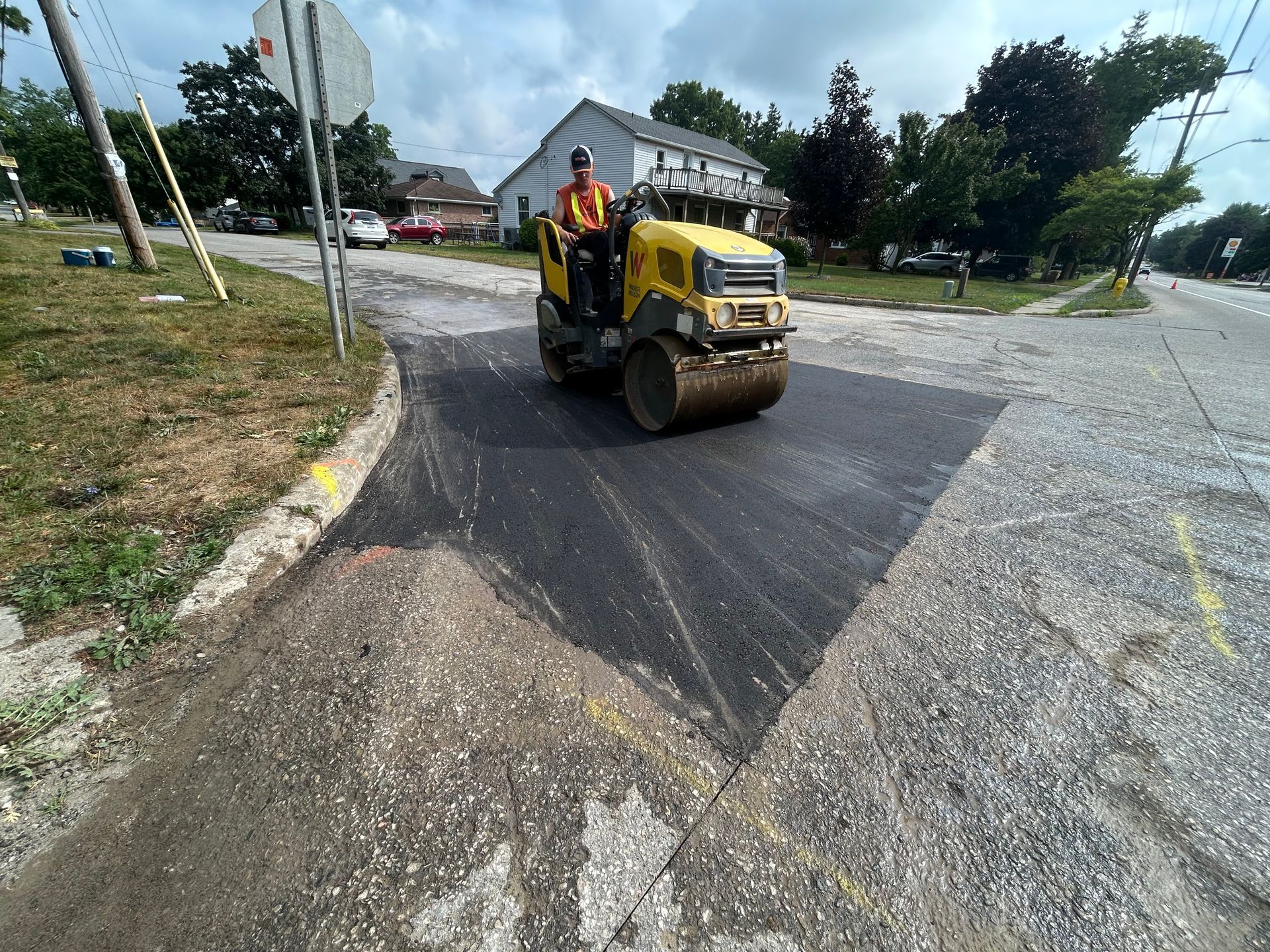 A worker operates a yellow asphalt roller to compact a fresh patch of road on a residential street.