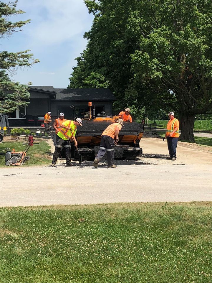 Construction workers in high-visibility vests operate a paving machine on a sunny residential road.