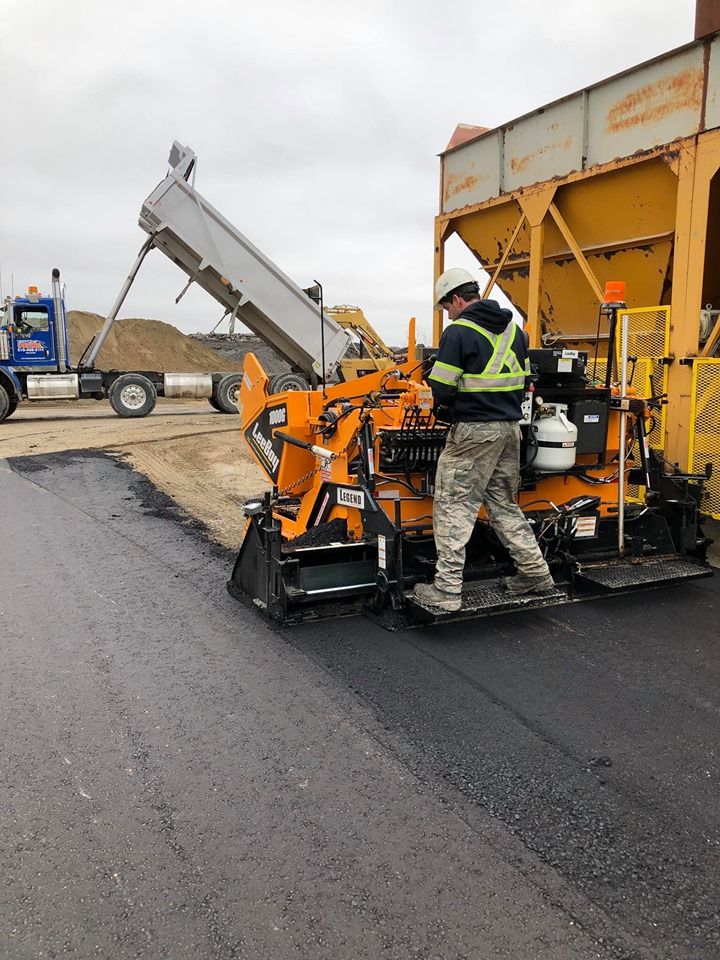 A worker in a high-visibility vest operates a paving machine as a dump truck unloads asphalt at a construction site.