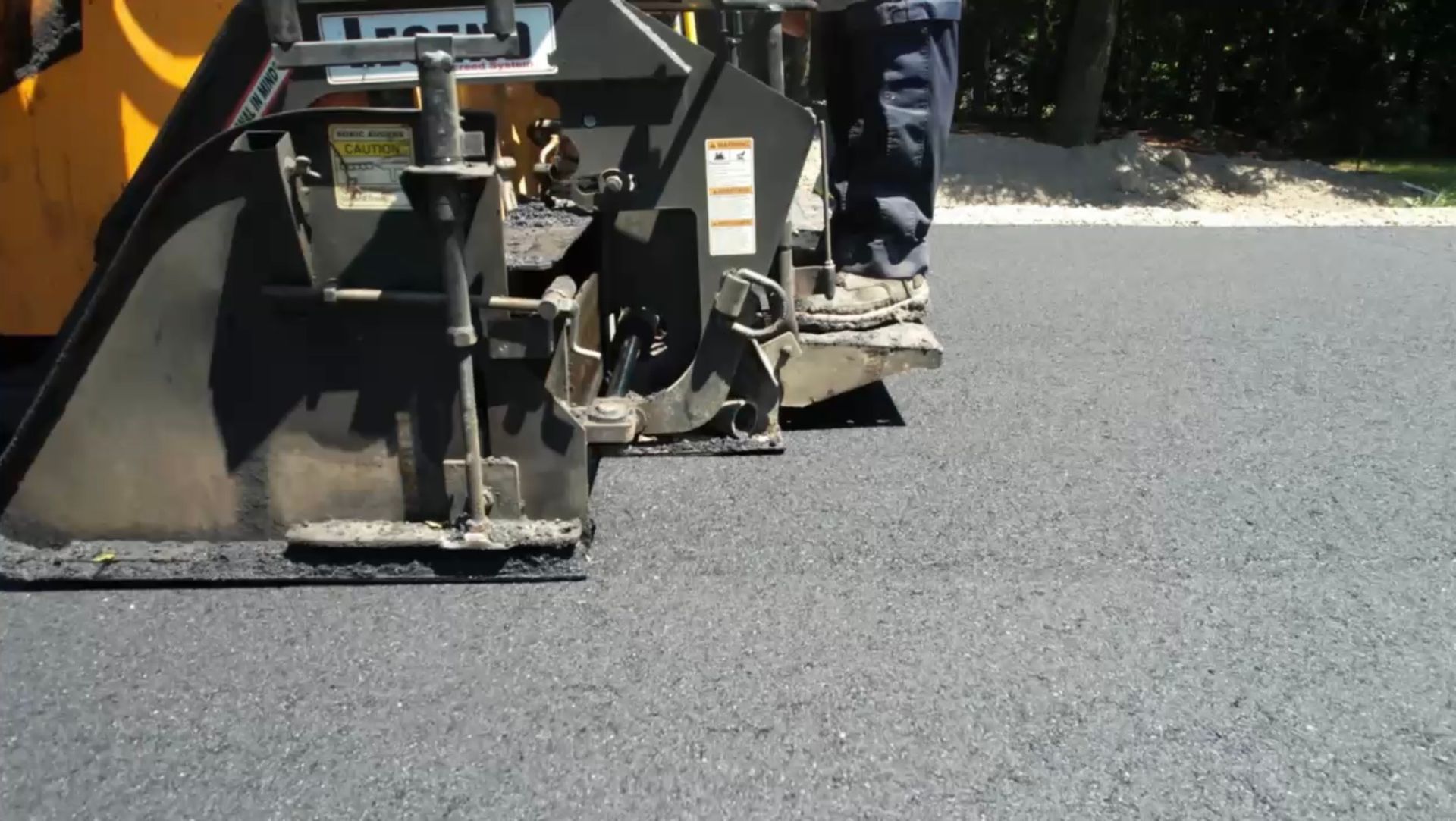 A paving machine lays fresh, dark asphalt on a road, with a worker standing on the equipment.