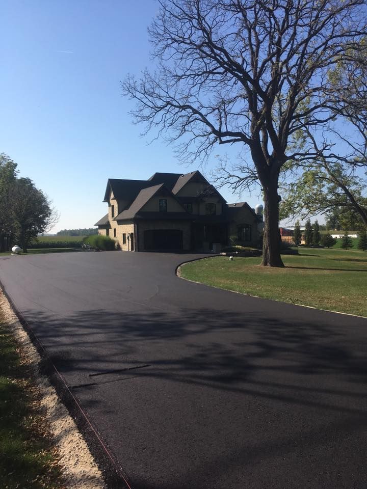 A freshly paved dark asphalt driveway leads toward a two-story suburban home on a sunny day.