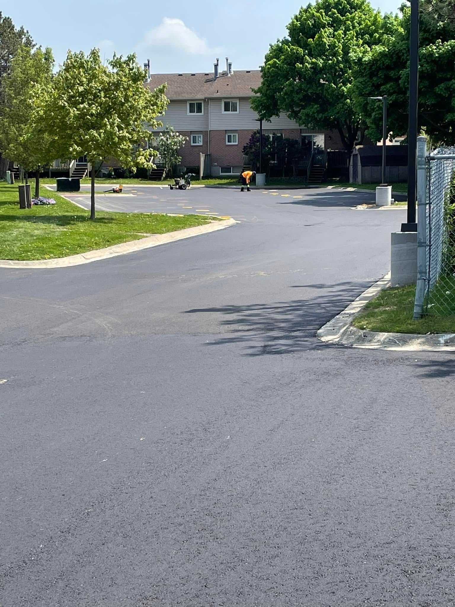 An asphalt parking lot area with trees and grass, leading to a multi-story residential building.