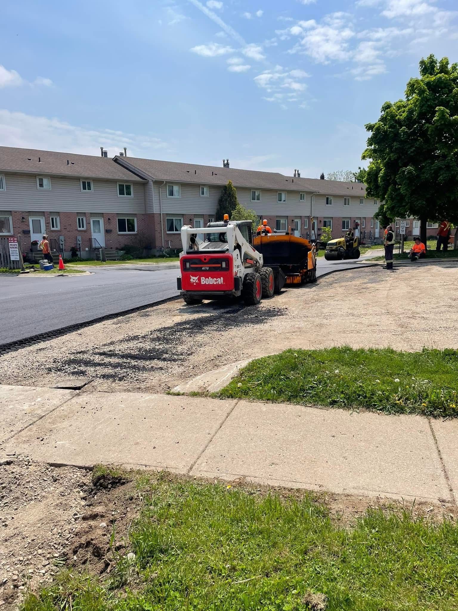 Construction workers operate a red Bobcat skid-steer loader to pave a gravel driveway in front of a townhouse complex.