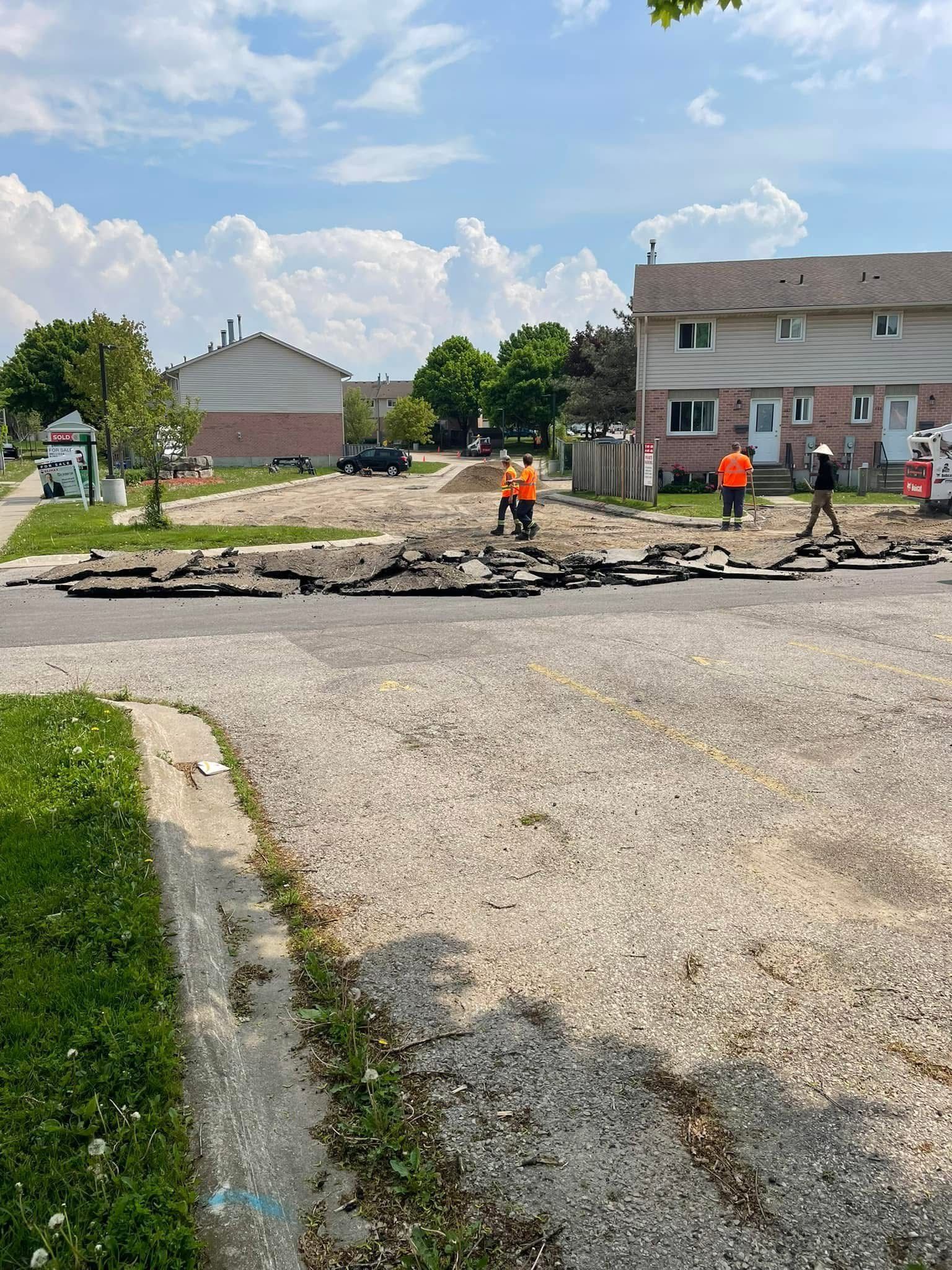 Construction workers in orange safety vests stand on a gravel road by a residential building under renovation.