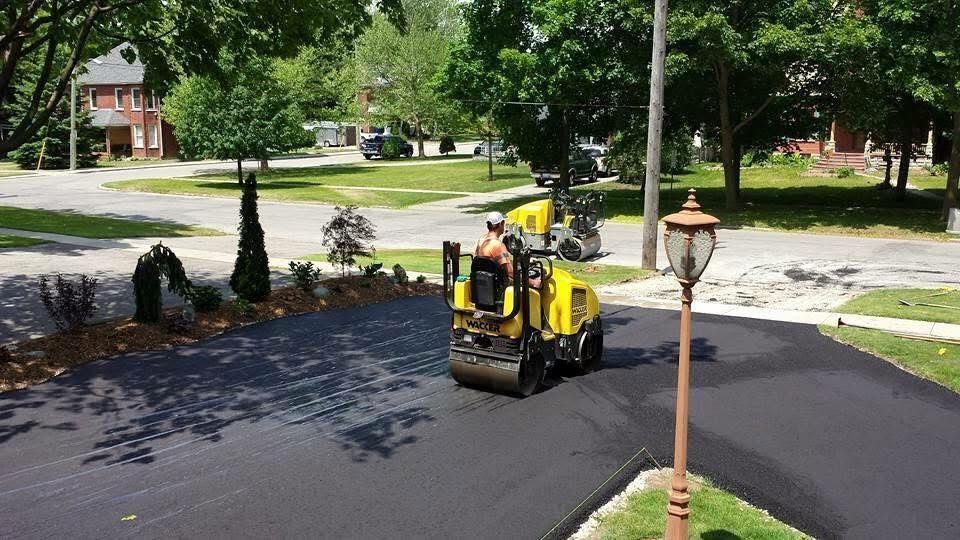 A yellow paving roller smooths a newly laid black asphalt driveway in a suburban neighborhood.