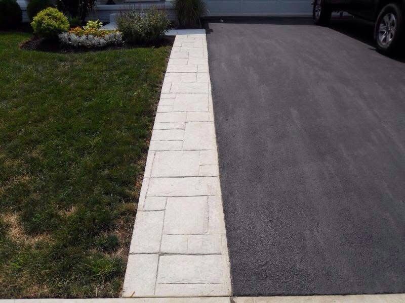 A white patterned concrete border separates a patch of green grass from a dark gray asphalt driveway.