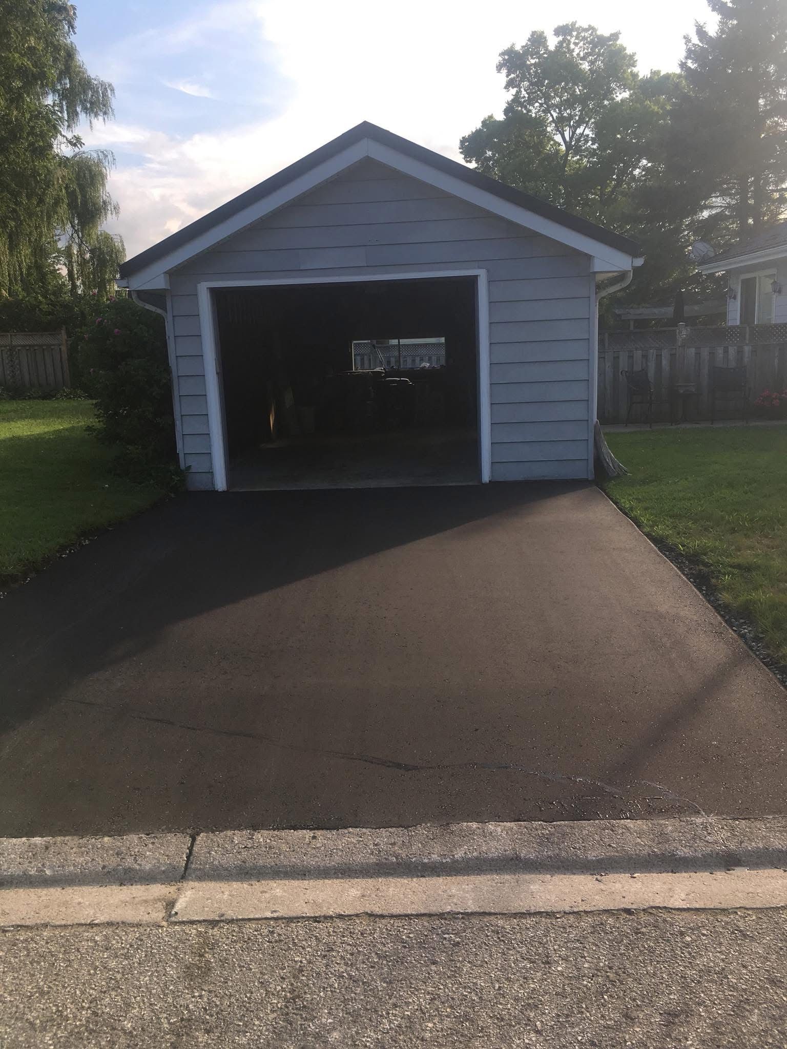 A gray detached garage with an open door and a newly paved black asphalt driveway.