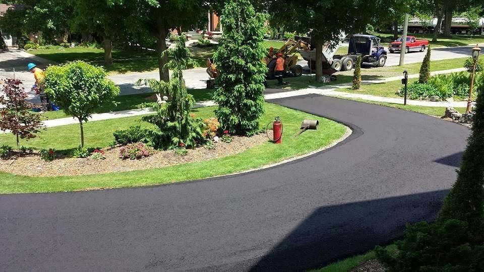 A freshly paved, curved asphalt driveway winds through a green front yard with landscaped trees and a fire hydrant.