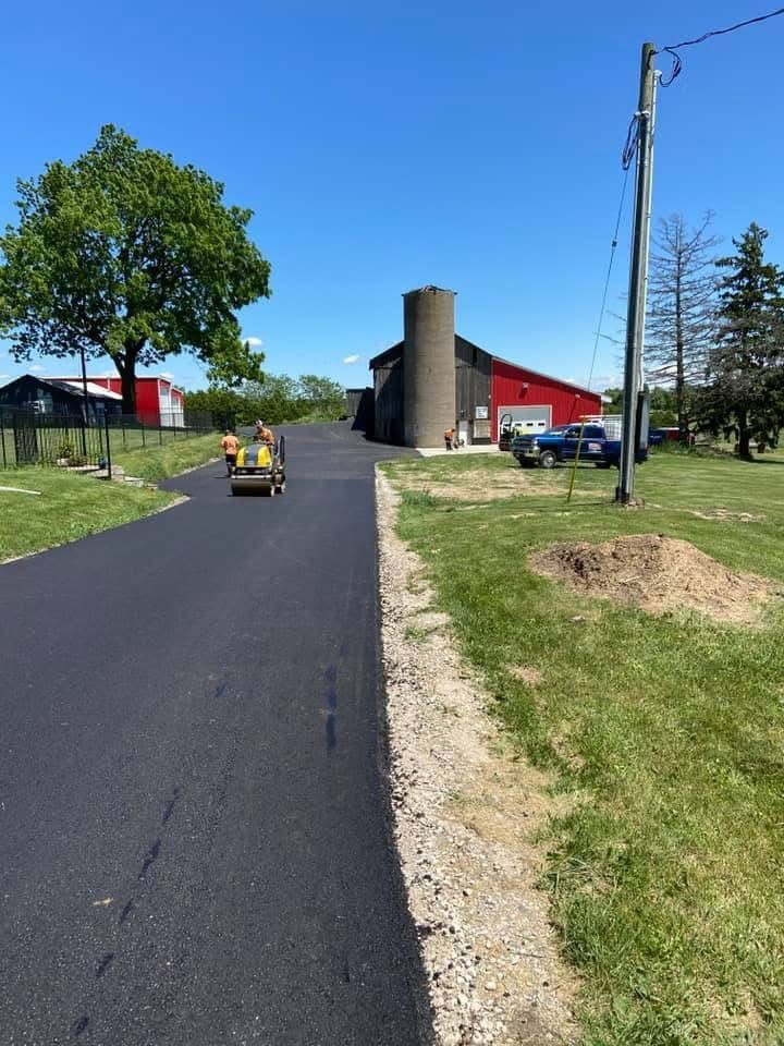 Workers operate a roller on a newly paved black asphalt driveway leading toward a red barn and silo on a sunny day.