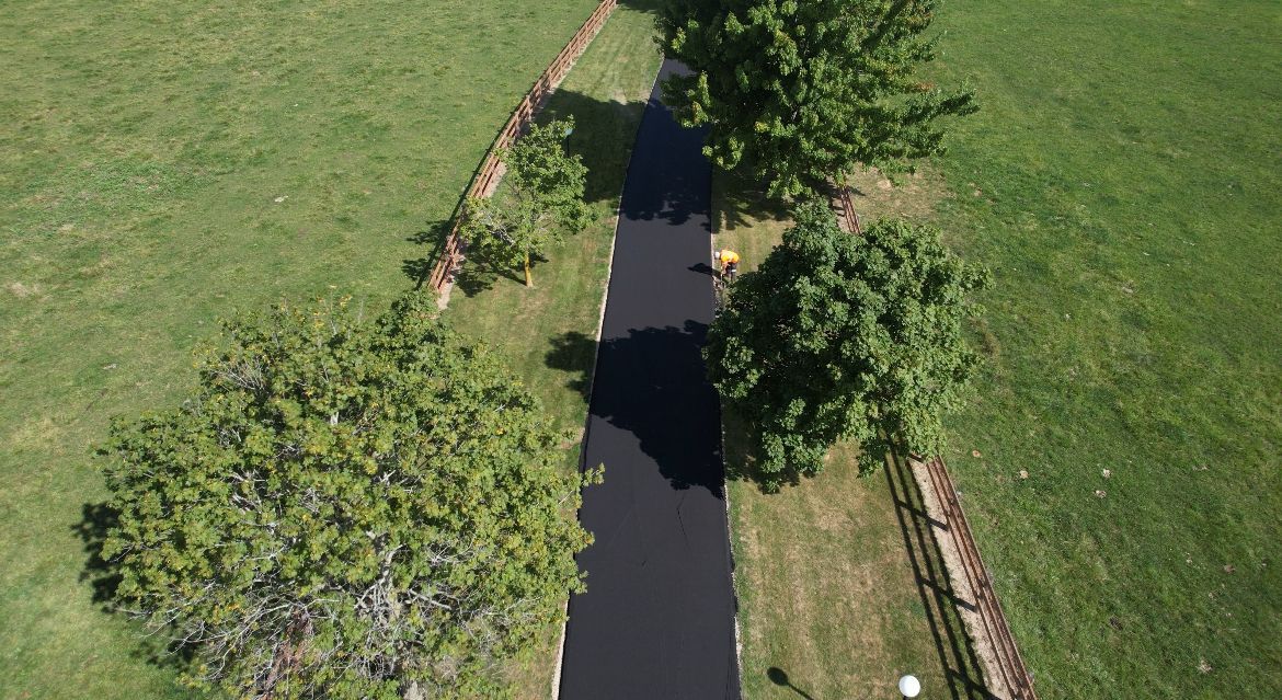 An aerial view of a dark asphalt pathway winding through a grassy lawn bordered by trees and a stone wall.