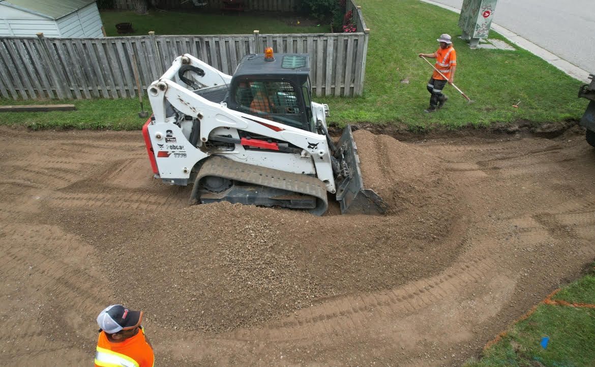 A white Bobcat skid-steer loader moves gravel on a construction site with two workers nearby on a grassy lawn.