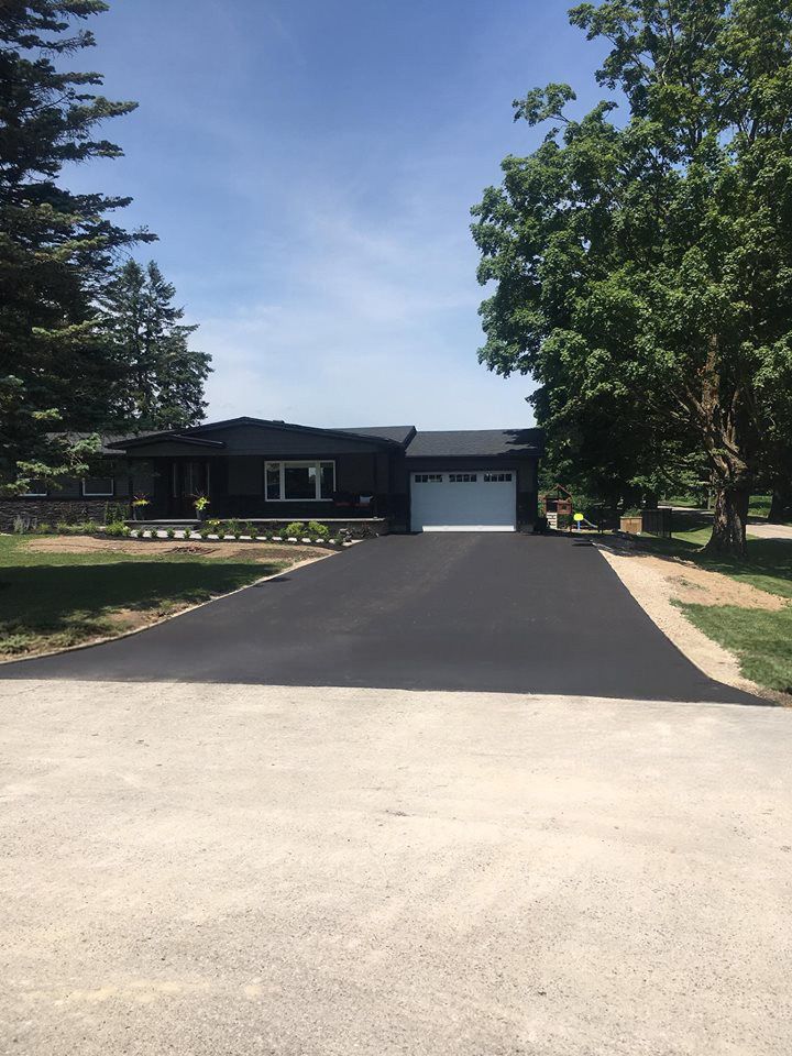 A paved driveway leading to a dark-sided one-story house with a white garage door, surrounded by green trees and grass.