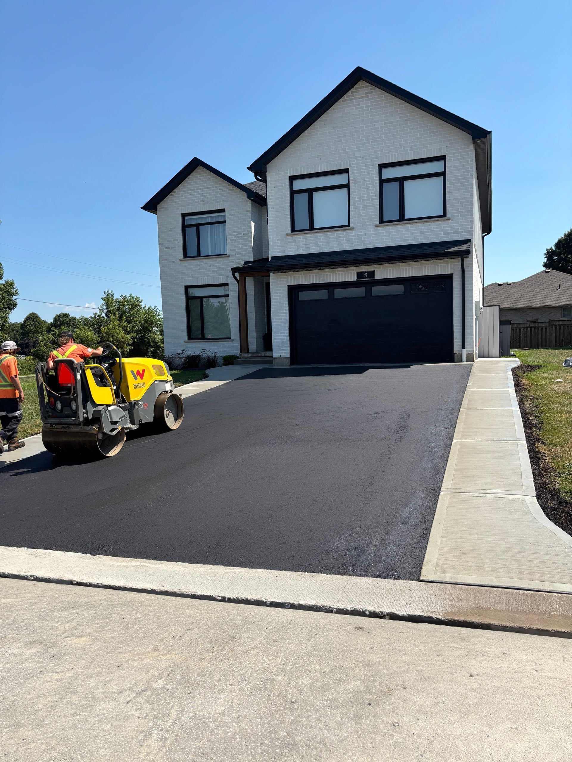 A construction worker operates a yellow asphalt roller on a newly paved driveway in front of a white two-story house.