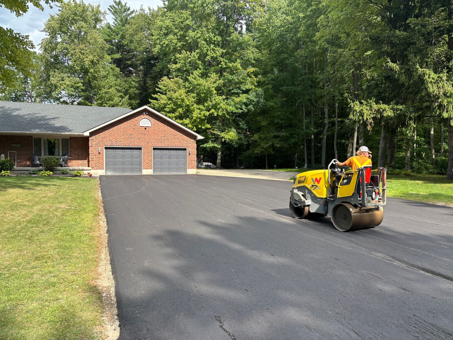A construction worker operates a yellow asphalt roller to smooth a newly paved driveway in front of a brick house.
