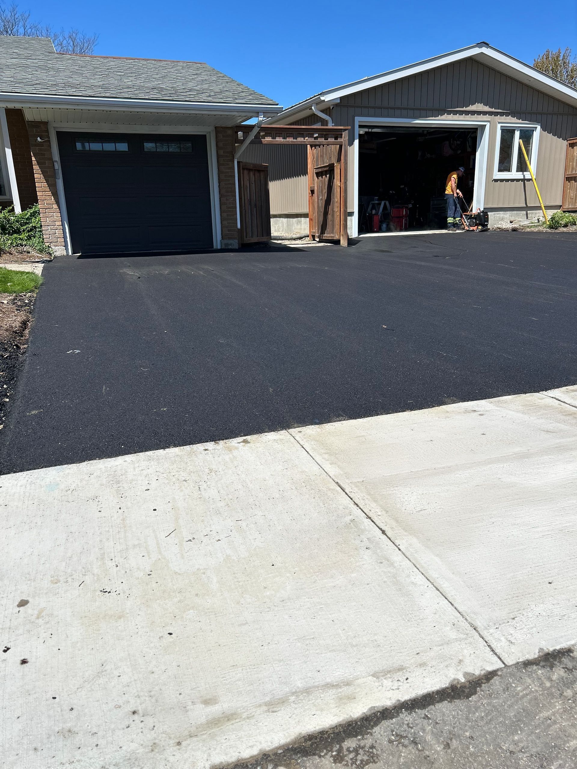 A freshly paved asphalt driveway leading to a two-car garage next to a concrete sidewalk under a clear blue sky.